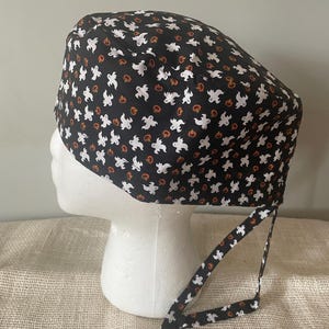 May include: Black scrub hat with a white ghost and orange pumpkin pattern. The hat has a tie on the back.