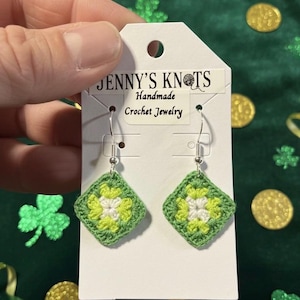 Op de afbeelding: Handgemaakte gehaakte oorbellen met een vierkant ontwerp. De oorbellen hebben een groene rand met een wit en lichtgroen bloemmotief in het midden. De oorbellen worden gepresenteerd op een witte kaart met de tekst "JENNY'S KNOTS Handmade Crochet Jewelry".