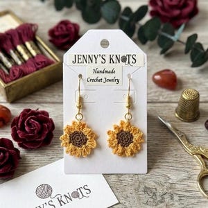 May include: A pair of handmade crochet sunflower earrings with gold-colored hooks. The earrings are yellow with a brown center, displayed on a white card with the text "JENNY'S KNOTS Handmade Crochet Jewelry".