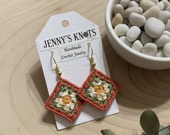 May include: A pair of handmade crochet earrings with a floral pattern. The earrings feature a peach border, green and white detailing, and a yellow centre. The earrings are displayed on a tag that reads "JENNY'S KNOTS Handmade Crochet Jewelry."