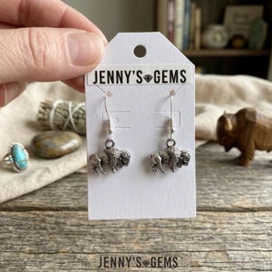 May include: Silver-toned buffalo earrings on a white card with the text "JENNY'S GEMS". The earrings are detailed with a textured buffalo design. The card is held against a wooden surface.