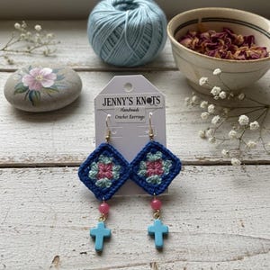 May include: Handmade crochet earrings with a blue, pink, and green floral design. The earrings have a square shape with a blue border, pink and green center, pink beads, and light blue cross charms. The earrings are labeled "Jenny's Knots".