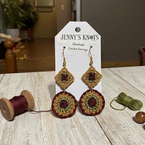 May include: Handmade crochet earrings with a geometric design. The earrings feature a square top and a circular bottom, with gold-colored hooks. The color scheme includes shades of brown, green, and burgundy. The tag reads "Jenny's Knots Handmade Crochet Earrings."