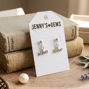 Może przedstawiać: Srebrne kolczyki z napisem "Love" w ozdobnej czcionce, prezentowane na białej karcie z napisem "Jenny's Gems". Kolczyki są umieszczone na tle starych książek i drewnianej powierzchni, z perłą i gałązkami lawendy.