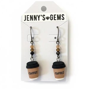 Op de afbeelding: Dangle oorbellen met miniatuur koffiekopjes. De bekers zijn bruin met zwarte deksels en het woord "Coffee" erop gedrukt. De oorbellen hebben een kralenaccent met bruine en zwarte kralen. Ze worden gepresenteerd op een witte kaart met de tekst "JENNY'S GEMS".