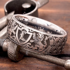 May include: A silver ring with an intricate design featuring a shield with a letter "L" inside, surrounded by lions and other decorative elements.