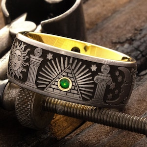 May include: A black and gold ring with an engraved design featuring a pyramid, an all-seeing eye, and other Masonic symbols. The ring has a green gemstone set in the center of the pyramid.