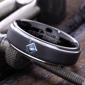 May include: A dark gray tungsten carbide ring with a blue gemstone set in the center. The ring is engraved with the words "and forever" on the inside of the band.