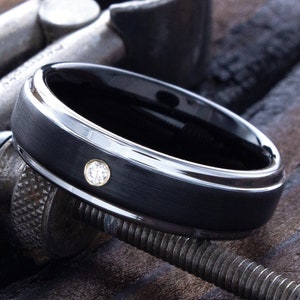 May include: A black ceramic ring with a single diamond set in the band. The ring is being measured with a metal tool.