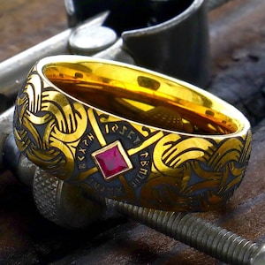 May include: A gold ring with a black and gold design and a red gemstone. The ring is in a metal clamp.