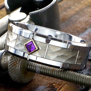 May include: A silver and white wedding band with a hammered finish and a square purple gemstone set in the center.