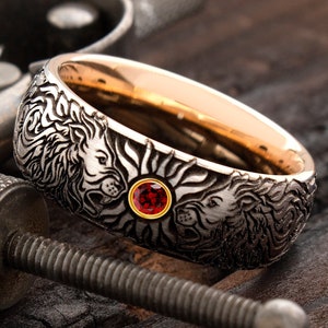 May include: A silver and gold ring with a red gemstone set in the center. The ring is engraved with two lions facing each other, with a sunburst design behind them.