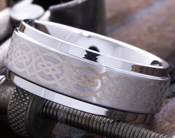 May include: A silver-colored ring with an engraved Celtic knot design. The ring is set on a metal clamp against a wooden surface, showcasing its intricate details and craftsmanship. The ring's polished surface reflects the light.