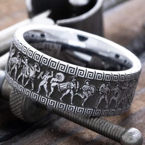 May include: A silver ring with a detailed engraved design of ancient Greek figures and a geometric border. The ring is displayed on a metal tool against a wooden surface, showcasing its intricate craftsmanship and historical theme.