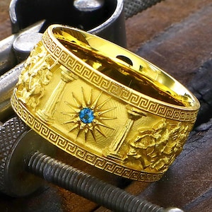 May include: A gold ring featuring a blue gemstone and intricate Greek-inspired designs. The ring is set against a wooden background.