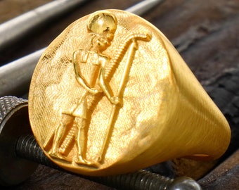 May include: A gold ring with a raised design of an Egyptian god holding a staff. The ring is on a wooden surface.