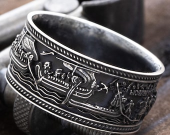 May include: A wide silver-toned metal ring featuring an embossed relief design of Viking longships sailing on stylized waves. The band has a dark, oxidized finish in the recessed areas to highlight the intricate nautical details.