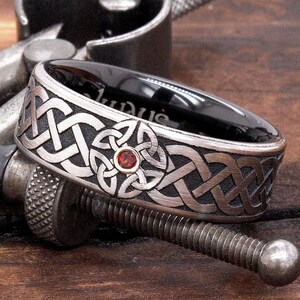May include: A black and silver ring with a Celtic knot design and a red gemstone in the centre. The ring is on a wooden surface.