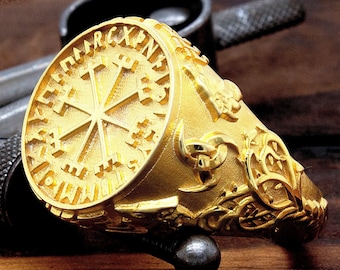 May include: A gold ring with a detailed design featuring a circular center with a symbol and runes around the edge. The ring is decorated with intricate carvings and patterns.