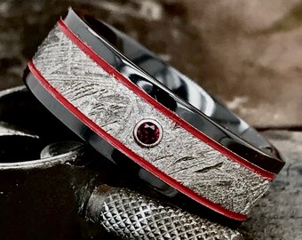 Peut inclure: Une bague noire et argentée avec une incrustation rouge et une petite pierre précieuse rouge. La bague est en métal et a une surface texturée.