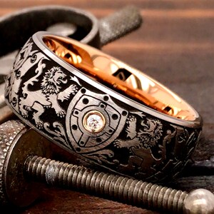 May include: A detailed ring with a black exterior featuring engraved lions and shields. The ring has a gold interior and a small diamond set in the center of a shield design. The ring is set on a wooden surface.
