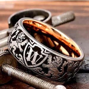 Puede incluir: Una banda de boda de plata y oro rosa con un intrincado diseño grabado que presenta leones y un escudo con la letra "W".