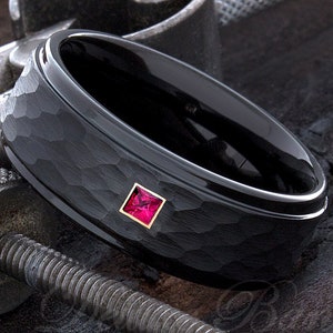 May include: A black tungsten carbide ring with a hammered finish and a small red gemstone set in a gold bezel.