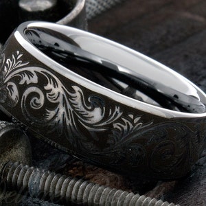 Puede incluir: Un anillo de metal gris oscuro con un interior de plata pulida y una superficie exterior grabada con intrincados patrones florales en espiral. La banda tiene un borde liso y redondeado y un alto contraste entre el exterior mate oscuro y la banda interior plateada brillante.