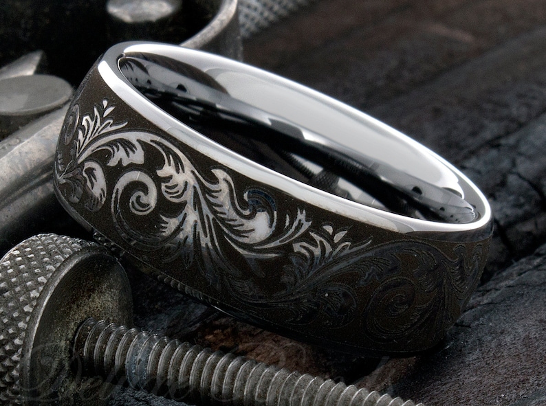 Puede incluir: Un anillo de metal gris oscuro con un interior de plata pulida y una superficie exterior grabada con intrincados patrones florales en espiral. La banda tiene un borde liso y redondeado y un alto contraste entre el exterior mate oscuro y la banda interior plateada brillante.
