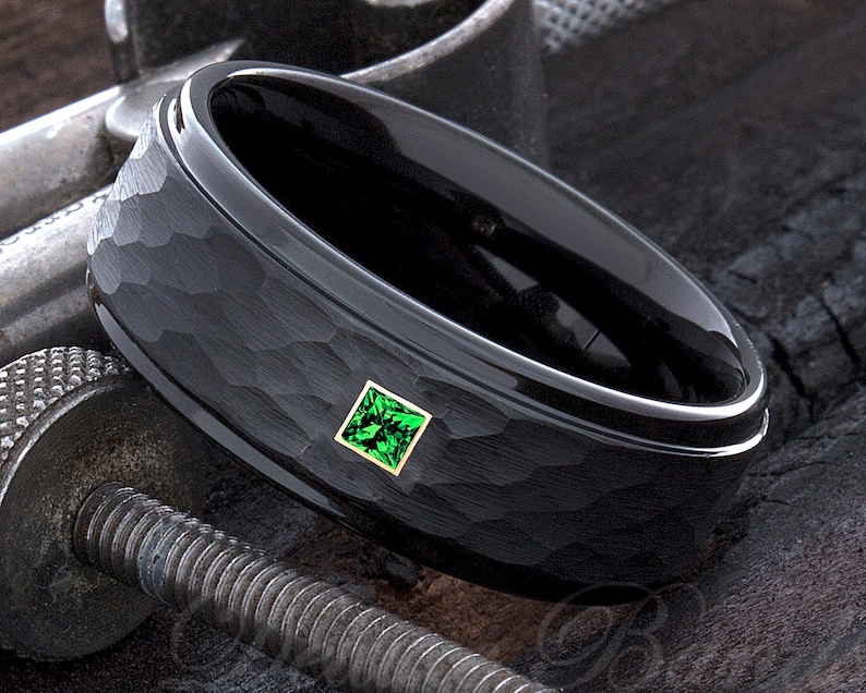 Op de afbeelding: Een zwarte ring met een gehamerde textuur en een vierkante, smaragdgroene edelsteen gezet in goud. De ring heeft een gepolijste, afgeschuinde rand. De ring wordt getoond op een metalen oppervlak.