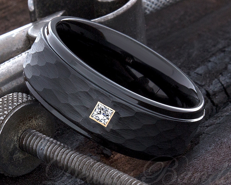 Op de afbeelding: Een zwarte ring met een getextureerd oppervlak en een vierkante, heldere edelsteen gezet in een goudkleurige bezel. De ring heeft een gepolijste binnenband en wordt getoond op een donker, getextureerd oppervlak.