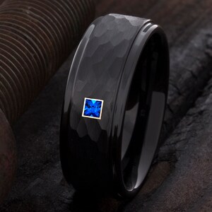 Op de afbeelding: Een zwarte ring met een gehamerde textuur en een vierkante, blauwe edelsteen gezet in een gouden frame. De ring heeft een gepolijste, glanzende afwerking en een modern design. De edelsteen is helderblauw.
