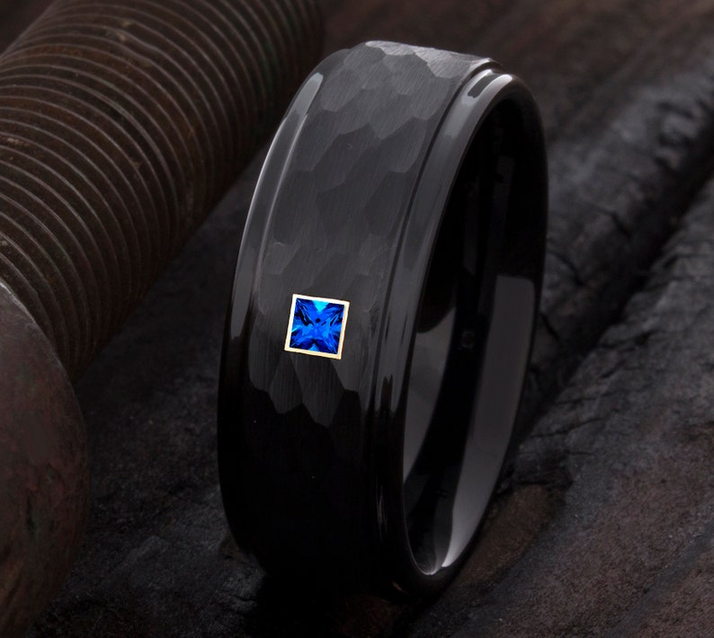 Op de afbeelding: Een zwarte ring met een gehamerde textuur en een vierkante, blauwe edelsteen gezet in een gouden frame. De ring heeft een gepolijste, glanzende afwerking en een modern design. De edelsteen is helderblauw.