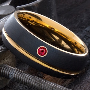 Puede incluir: Un anillo de carburo de tungsteno negro con una banda dorada y una pequeña piedra preciosa roja engastada en el centro.