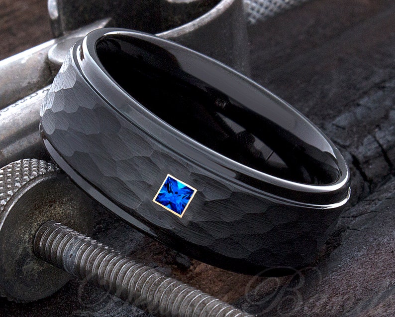 Op de afbeelding: Een zwarte ring met een gehamerde textuur en een vierkante, blauwe edelsteen in een gouden montuur. De ring heeft een gepolijste binnenband en ligt op een metalen oppervlak.