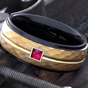 May include: A black and gold men's ring with a hammered finish and a red gemstone in the center.
