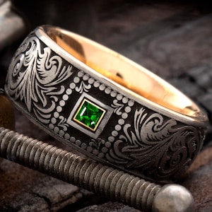 May include: A black and gold wedding band with an intricate engraved design and a green gemstone set in the center.
