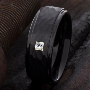 Op de afbeelding: Zwarte ring met een gehamerde textuur en een vierkante, heldere edelsteen gezet in een goudkleurige bezel. De ring heeft een gepolijste, glanzende afwerking op de binnen- en buitenranden. Close-up.