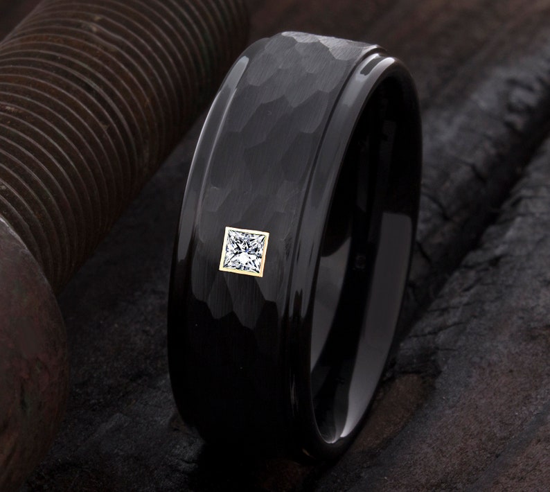 Op de afbeelding: Zwarte ring met een gehamerde textuur en een vierkante, heldere edelsteen gezet in een goudkleurige bezel. De ring heeft een gepolijste, glanzende afwerking op de binnen- en buitenranden. Close-up.