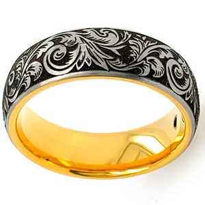 Puede incluir: Un anillo con una banda interior de color dorado pulido y una superficie exterior de color gris oscuro. La banda exterior est&aacute; decorada con un detallado patr&oacute;n de volutas grabadas en tono plateado que rodea toda la circunferencia.
