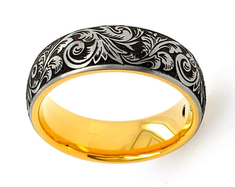 Puede incluir: Un anillo con una banda interior de color dorado pulido y una superficie exterior de color gris oscuro. La banda exterior est&aacute; decorada con un detallado patr&oacute;n de volutas grabadas en tono plateado que rodea toda la circunferencia.