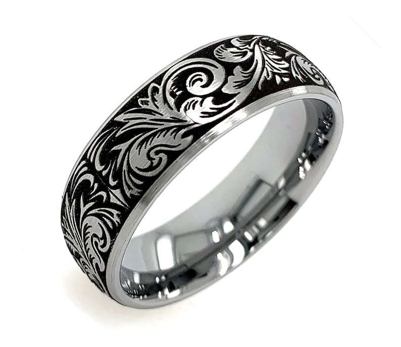 Puede incluir: Un anillo de metal plateado con un acabado pulido en el interior y una incrustaci&oacute;n negra en contraste en el exterior. La superficie negra est&aacute; grabada con un patr&oacute;n continuo de volutas florales de color plateado alrededor de la banda.
