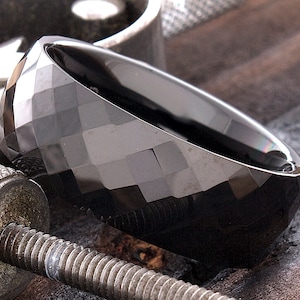 May include: A black tungsten carbide ring with a faceted, geometric design. The ring is shown on a textured surface.