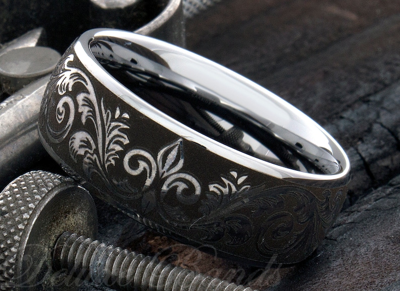 Puede incluir: Un anillo de metal gris oscuro con un interior plateado pulido y una superficie exterior mate grabada con un detallado patr&oacute;n de volutas florales. La banda tiene un perfil ancho con un borde liso y redondeado y un acabado met&aacute;lico.