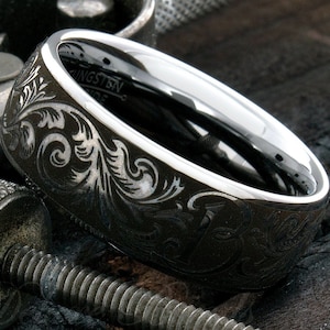 Puede incluir: Un anillo de carburo de tungsteno gris oscuro con un interior plateado pulido y una superficie exterior grabada con intrincados patrones florales en espiral. La palabra Tungsten es visible en la banda interior del anillo.