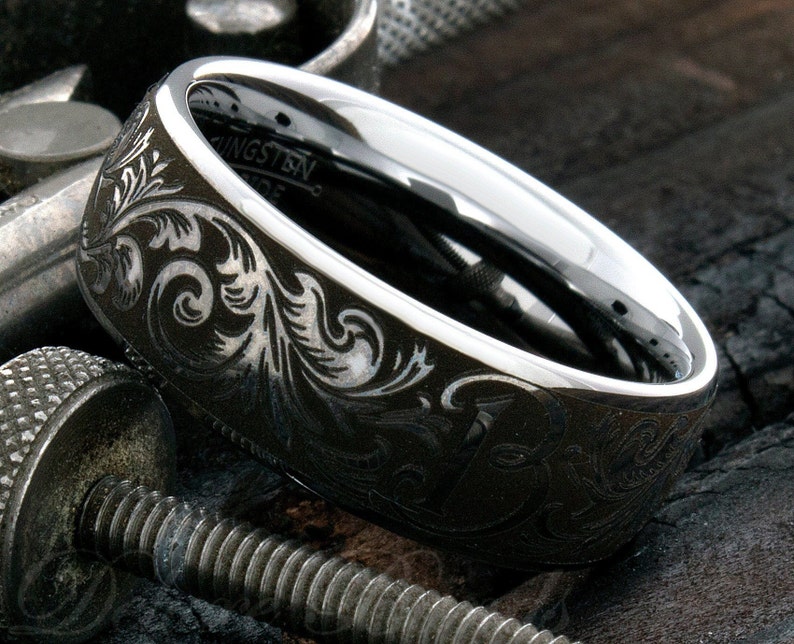 Puede incluir: Un anillo de carburo de tungsteno gris oscuro con un interior plateado pulido y una superficie exterior grabada con intrincados patrones florales en espiral. La palabra Tungsten es visible en la banda interior del anillo.