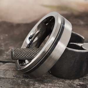 Puede incluir: Primer plano de dos anillos plateados. Uno tiene un acabado de metal cepillado con una fina banda negra y un detalle similar a una cuerda. El otro anillo es de metal mate más oscuro. Una herramienta de metal texturizada descansa entre los anillos.