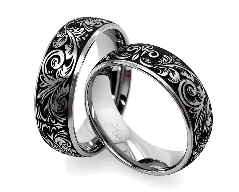 Puede incluir: Dos anillos de tono plateado con una incrustaci&oacute;n negra y un intrincado patr&oacute;n floral grabado. El interior de los anillos es de plata pulida e incluye el texto Tungsten Carbide grabado en la superficie interior de la banda.