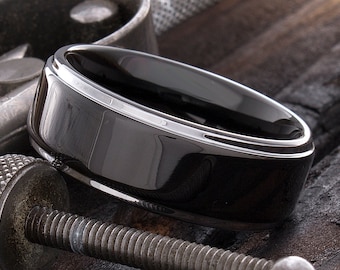May include: A black ceramic wedding band with a polished silver edge. The ring is shown on a textured surface.