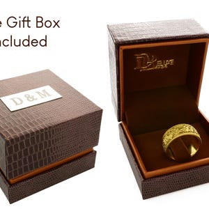 Puede incluir: Un anillo dorado con un patr&oacute;n texturizado intrincado se encuentra dentro de un joyero marr&oacute;n con relieve de cocodrilo. La tapa presenta una placa rectangular plateada con el texto D & M, mientras que el interior muestra el texto Deluxe.